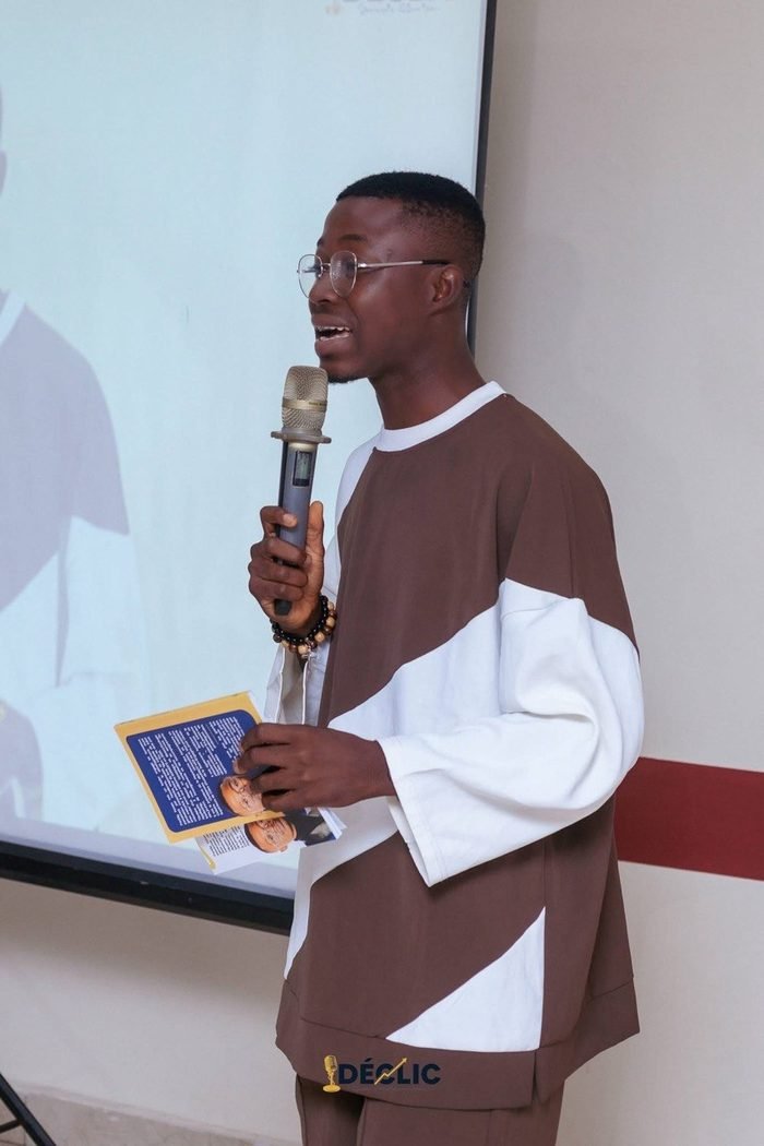 Freddy Konan au micro devant un écran de projection lors d’une conférence à Bouaké, une brochure à la main